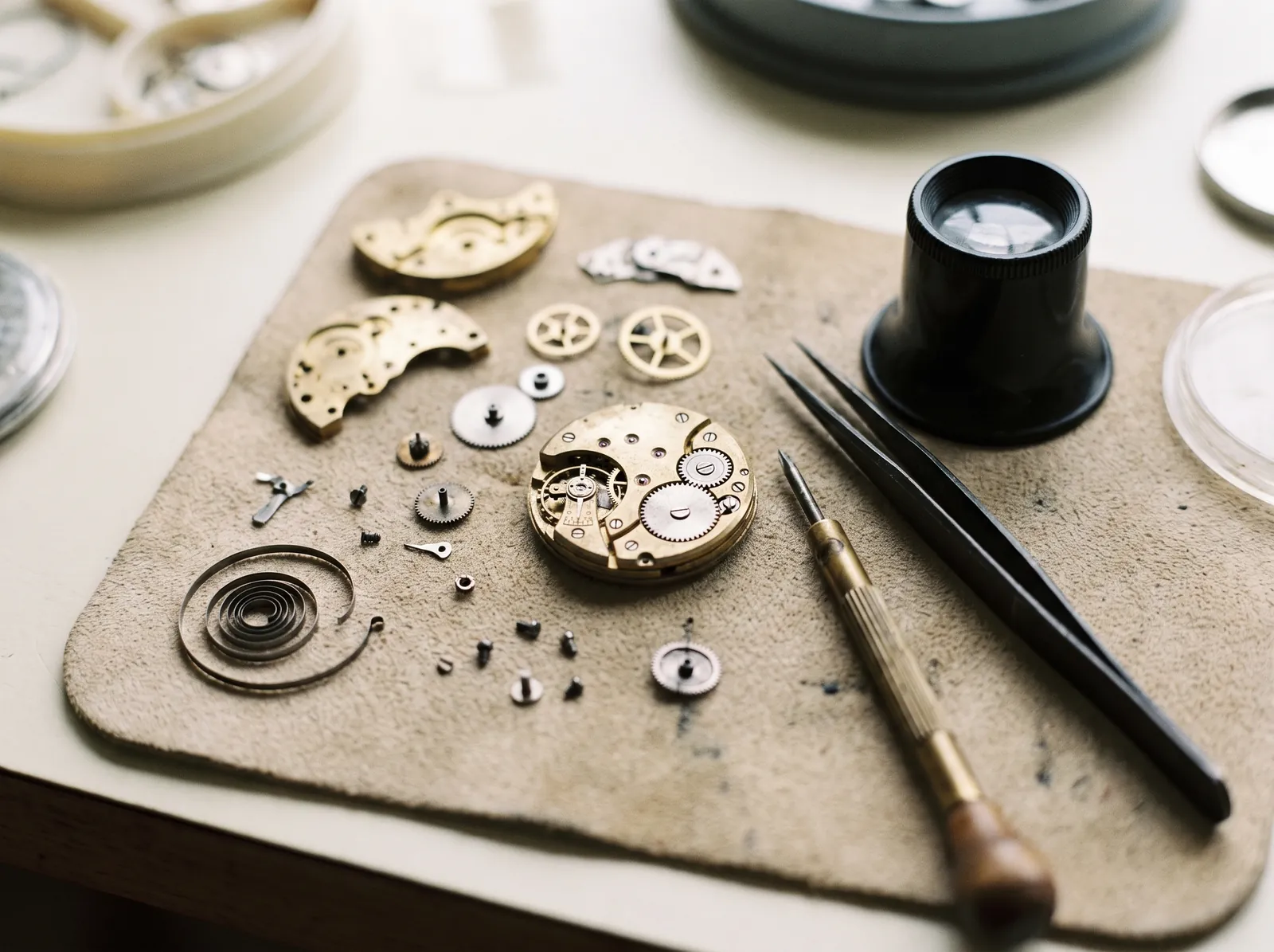 A watchmaker's bench from above: brass plates, wheels, tweezers, a loupe and a screwdriver on a suede pad